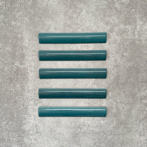Flat lay of Zellige Peacock Green 3×20cm Moroccan-style ceramic bullnose tiles, showcasing the rounded edge profile, deep blue-green glaze and subtle tonal variation on a neutral background.