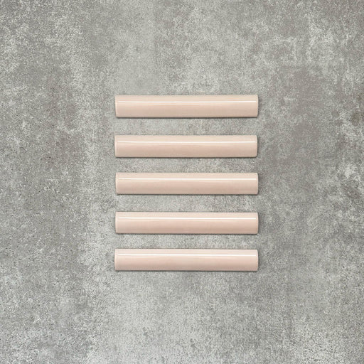 Flat lay of Zellige Pink Mist Bullnose 3 × 20cm Moroccan-style ceramic trim tiles in soft pale pink tones, displayed with a smooth rounded edge and glossy glaze, arranged on a textured grey background to highlight the handcrafted zellige-effect finish and decorative edging profile.