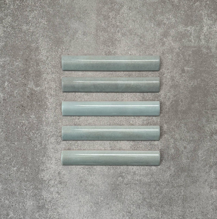 Flat lay of Zellige Aqua 3×20cm Moroccan-style ceramic bullnose tiles, showcasing the rounded edge profile, glossy blue-green glaze and subtle tonal variation on a neutral background.