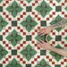 Close-up of Abstract Compass Green 20x20cm patterned tiles arranged to showcase the repeating geometric design in green, white and red tones, with hands demonstrating tile placement and alignment.
