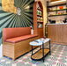 Café interior featuring Abstract Serit Patterned Tiles in a 20x20cm format, laid as a bold geometric floor design. The playful pattern combines soft pink semicircles with deep green striped detailing on a white background, paired with warm wooden furniture and blush seating to create a contemporary, characterful commercial space.