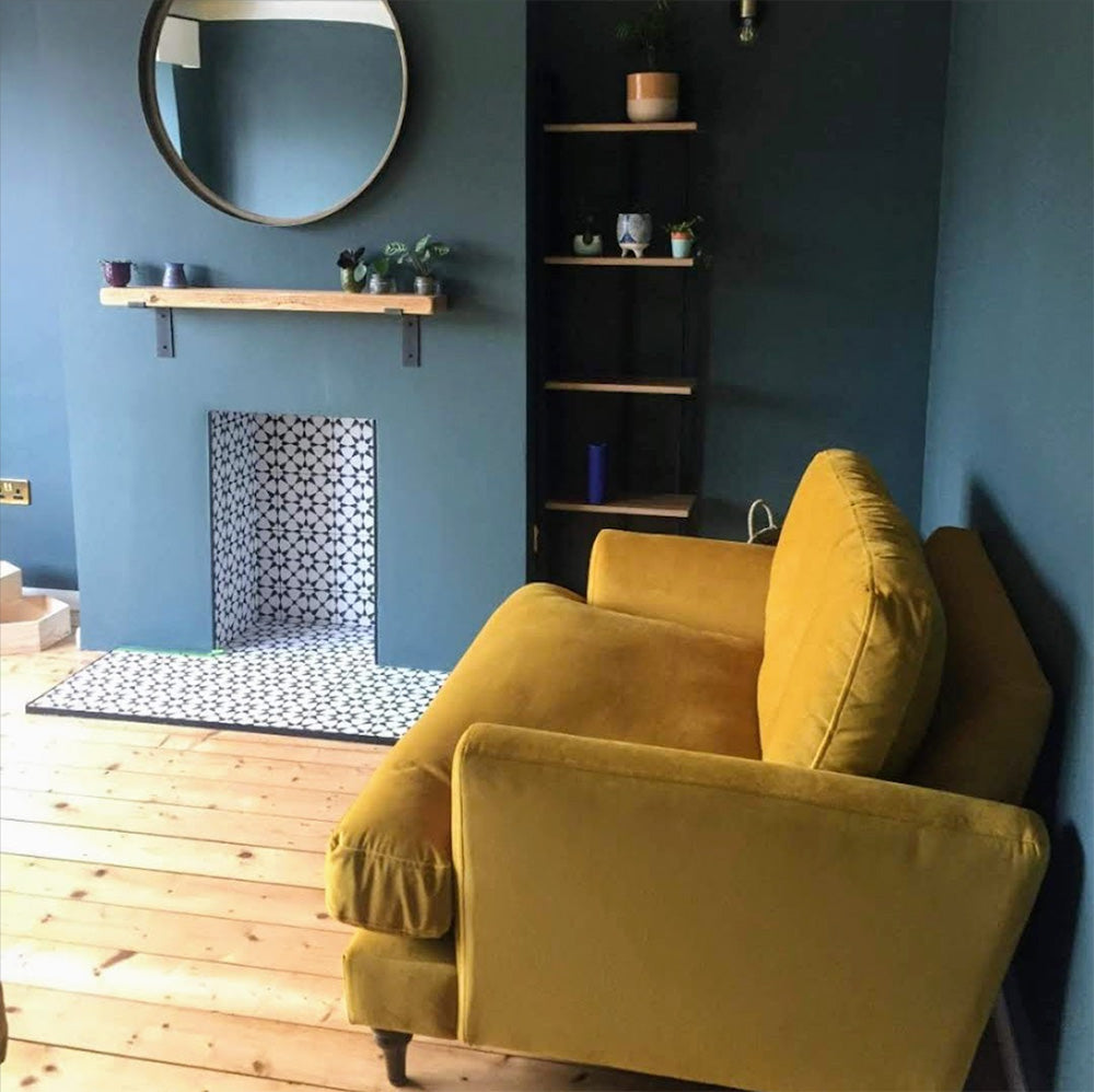 Living room featuring Agadir Black & White Mini Moroccan-style 15x15cm porcelain tiles used in a fireplace setting, showcasing a bold geometric star pattern, styled with deep blue walls, wooden floors and a mustard sofa.