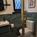 Bathroom interior featuring Allegra Green Gloss Brick ceramic tiles in a 6 × 25cm format, laid horizontally on the walls to create a rich green, glossy backdrop with subtle tonal variation, paired with brass fixtures and black-and-white patterned floor tiles.