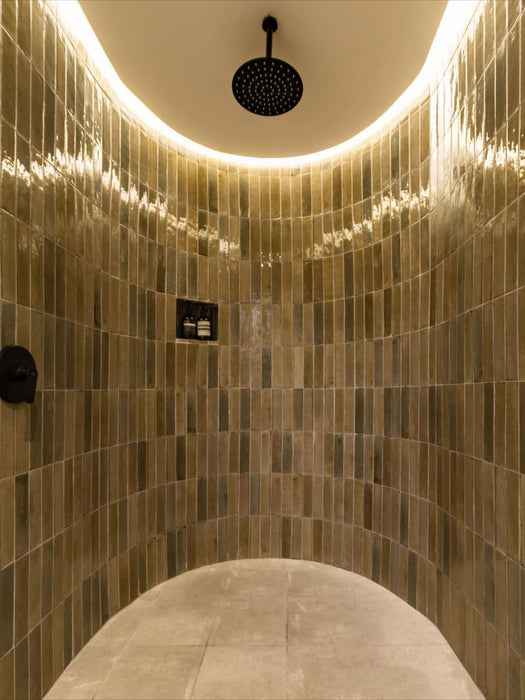 Curved shower enclosure lined with Allegra Sage Green gloss brick tiles in a vertical layout, highlighting a high-shine glazed finish with natural tonal variation, complemented by a black rainfall shower and soft integrated ceiling lighting.