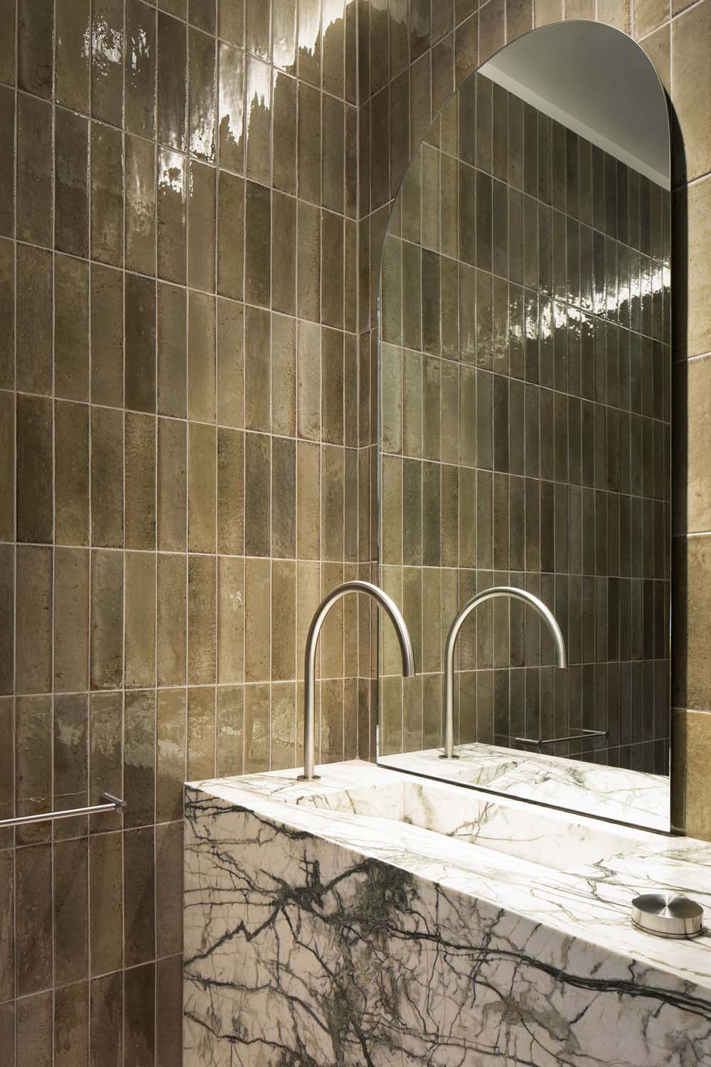 Bathroom wall finished with Allegra Sage Green gloss brick tiles in a vertical layout, featuring subtle tonal variation and a reflective glazed surface, paired with a marble vanity, wall-mounted basin and contemporary chrome tapware.