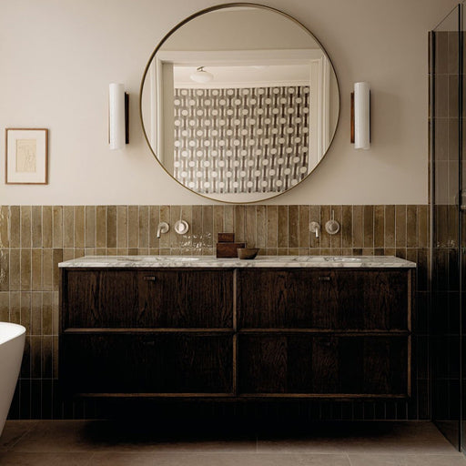 Allegra Sage Green 6x25cm gloss brick tiles installed as a bathroom feature wall, showcasing soft earthy green tones with gentle shade variation and a reflective glazed finish. The elongated format adds subtle texture and depth, complementing dark wood cabinetry, marble surfaces and warm brass accents for a refined, contemporary look.