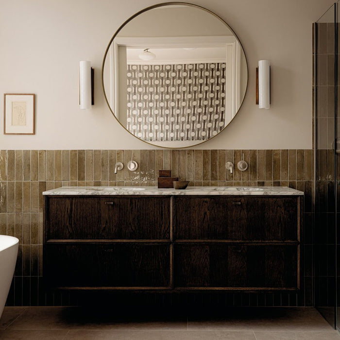 Allegra Sage Green 6x25cm gloss brick tiles installed as a bathroom feature wall, showcasing soft earthy green tones with gentle shade variation and a reflective glazed finish. The elongated format adds subtle texture and depth, complementing dark wood cabinetry, marble surfaces and warm brass accents for a refined, contemporary look.