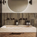 Allegra Sage Green 6x25cm gloss brick tiles used as a bathroom splashback, displaying rich sage tones with natural variation and a softly reflective glazed finish. Laid vertically to enhance height and texture, the handmade-effect surface pairs effortlessly with warm wood cabinetry, stone basins and dark bronze fittings for a contemporary, spa-inspired look.