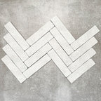 Product image of Amalfi Brick Carrara Vintage Marble wall and floor tiles in a 5.4cm x 22cm format, arranged in a herringbone pattern, showcasing a soft white marble effect surface with subtle tonal variation.