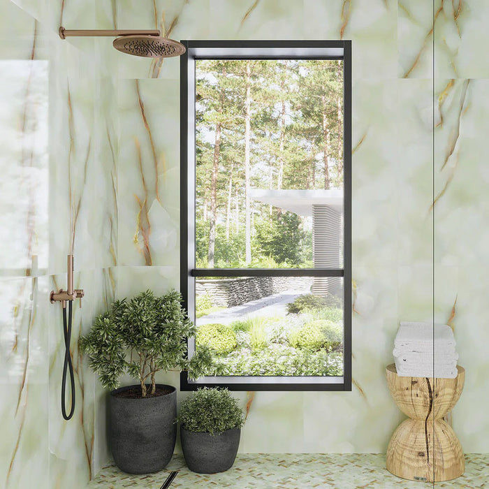 Bathroom interior featuring Anastasia Jade Green Onyx Matt 60 × 120cm stone effect wall tiles, with soft green tones and flowing gold veining, paired with brass shower fittings, indoor plants, and a large window.