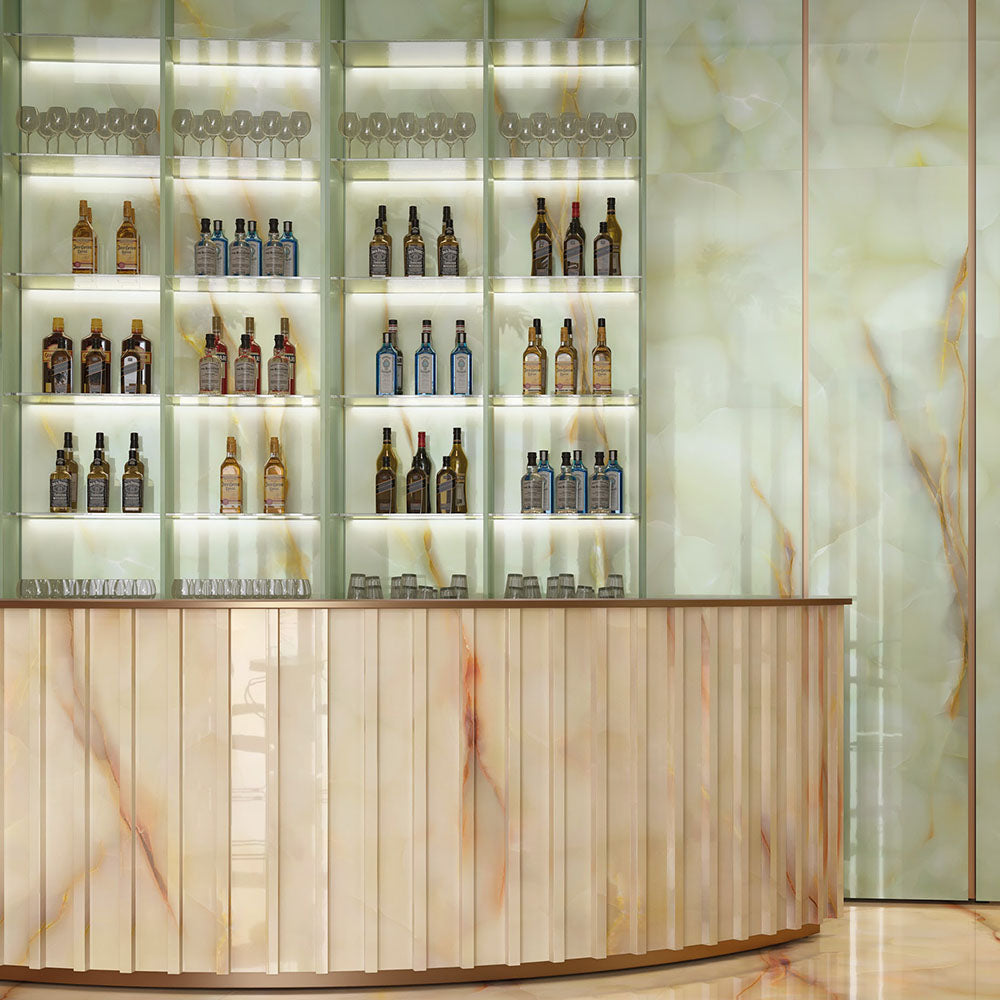 Bar interior featuring Anastasia Jade Green Onyx Polished 60 × 120cm stone effect wall and floor tiles, with soft green tones and flowing gold veining, paired with a curved bar, illuminated shelving, and warm metallic accents.