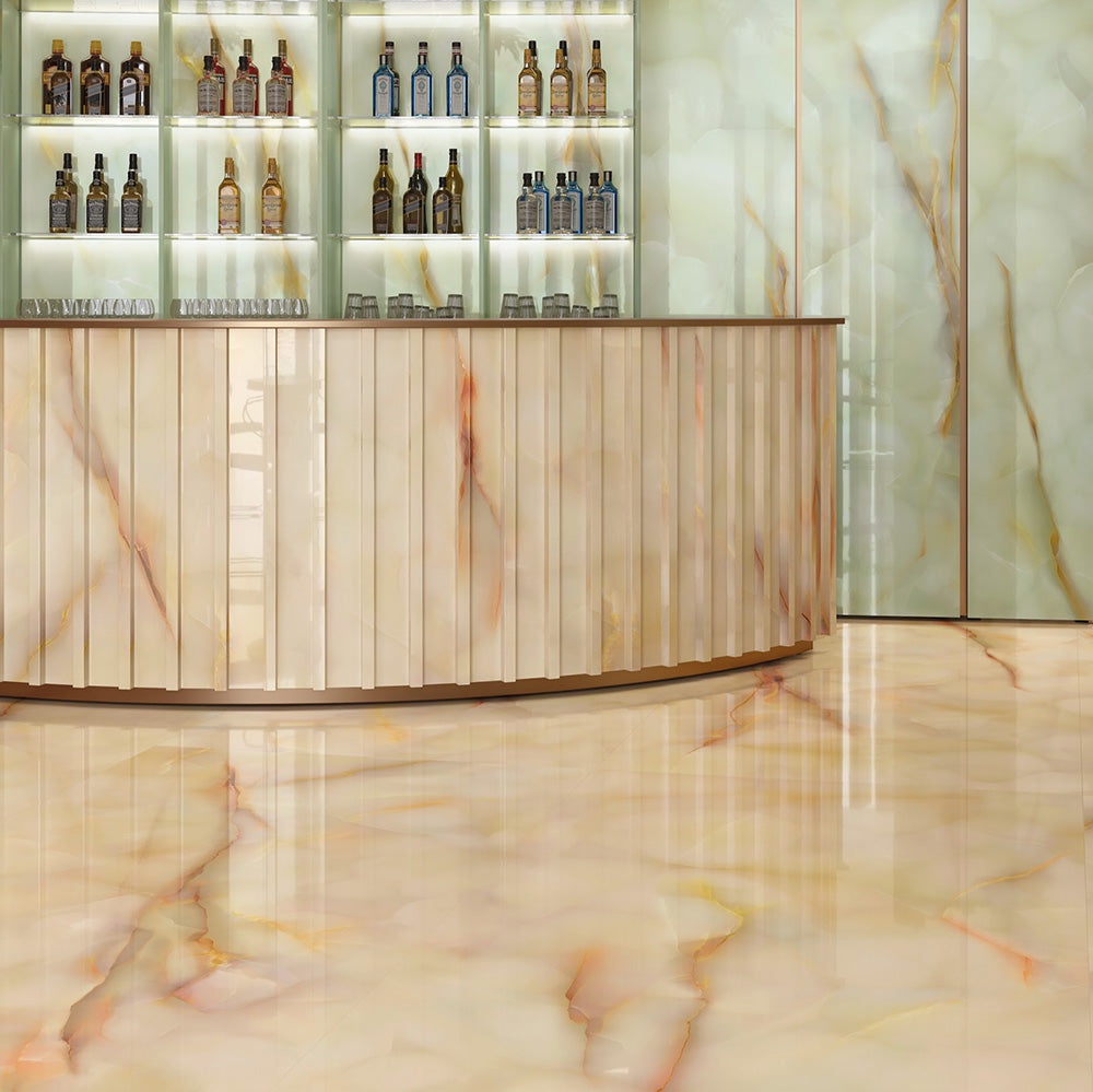 Bar interior featuring Anastasia Miele Onyx Polished 60 × 120cm stone effect floor tiles, with warm honey tones and flowing veining, paired with a curved bar, illuminated shelving, and soft green wall panels.