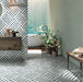 Bathroom interior featuring Art Deco Emerald Green marble effect patterned porcelain tiles 44 × 44cm, used across both wall and floor to create a continuous geometric layout. The bold linear pattern in deep green and white tones adds structure and movement, paired with a freestanding bath, wooden vanity unit, and natural greenery for a balanced, contemporary feel.
