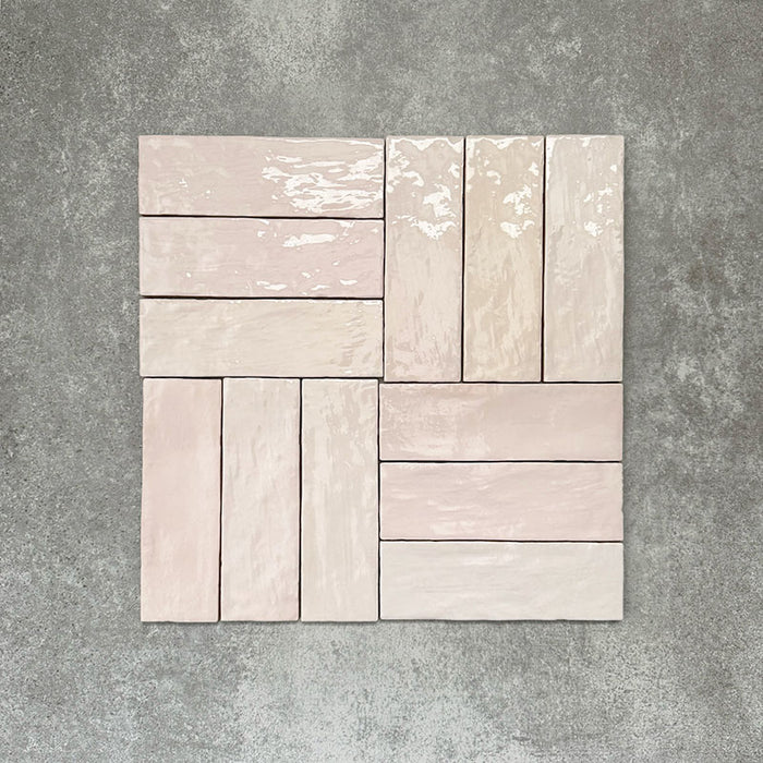 Flat lay of Zellige Pink Mist Brick 6.5 × 20cm Moroccan-style ceramic tiles in soft pale pink and blush tones, arranged in a basketweave layout on a textured grey background, showcasing the glossy glaze, gentle tonal variation and handcrafted zellige-effect finish.