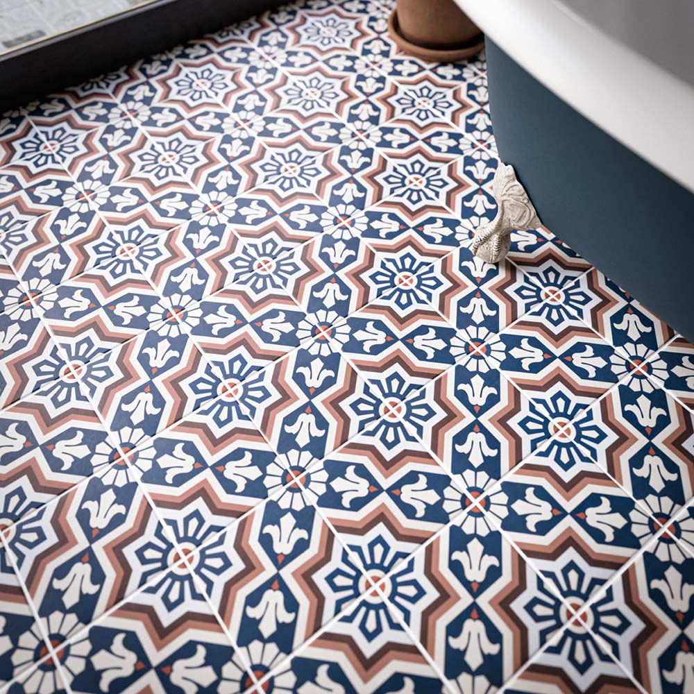 Close-up of Balthazar Grand 20x20cm Victorian and Moroccan-style patterned bathroom floor tiles, featuring intricate geometric and floral motifs in navy blue, terracotta and white, beside a freestanding bathtub.