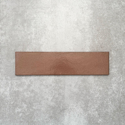 Baroque Cotto porcelain wall and floor tile in a 9.1cm x 37.5cm format, featuring a warm terracotta metallic finish with gently distressed edges, displayed against a textured concrete-effect background.