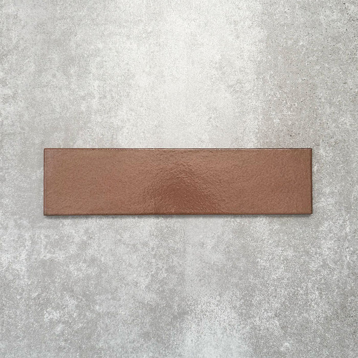 Baroque Cotto porcelain wall and floor tile in a 9.1cm x 37.5cm format, featuring a warm terracotta metallic finish with gently distressed edges, displayed against a textured concrete-effect background.