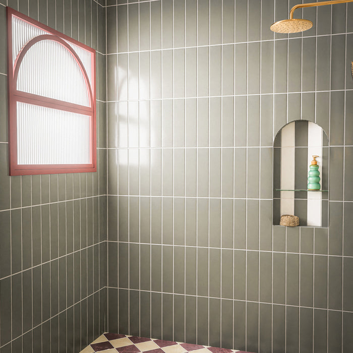 Contemporary walk-in shower featuring Baroque Lichen Green porcelain wall tiles in a vertical stacked layout, paired with a burgundy and white chequerboard floor, brushed brass shower fittings, an arched recessed niche and a statement red-framed window for a bold, modern bathroom design.