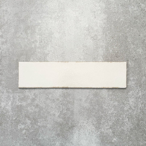 Baroque Vanilla porcelain wall and floor tile in a 9.1cm x 37.5cm format, featuring a soft cream metallic finish with gently distressed edges, displayed against a textured concrete-effect background.