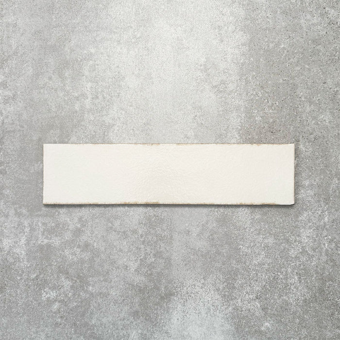 Baroque Vanilla porcelain wall and floor tile in a 9.1cm x 37.5cm format, featuring a soft cream metallic finish with gently distressed edges, displayed against a textured concrete-effect background.