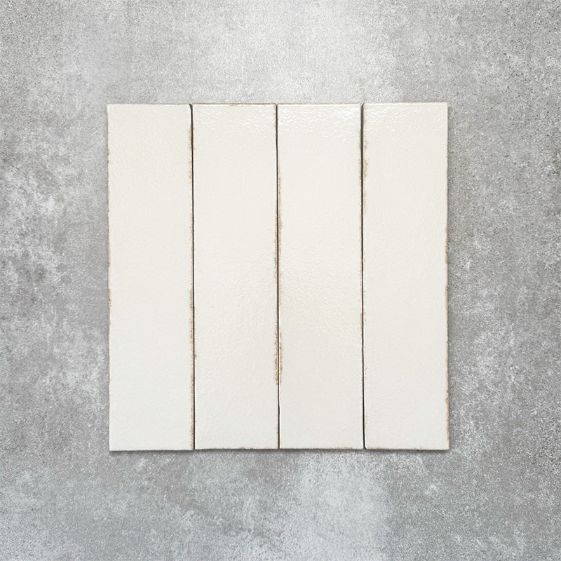 Baroque Vanilla porcelain wall and floor tile in a 9.1cm x 37.5cm format, featuring a soft cream metallic finish with gently distressed edges, displayed against a textured concrete-effect background.
