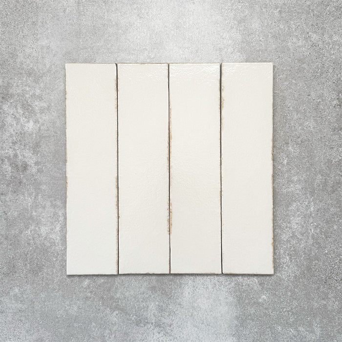 Baroque Vanilla porcelain wall and floor tile in a 9.1cm x 37.5cm format, featuring a soft cream metallic finish with gently distressed edges, displayed against a textured concrete-effect background.