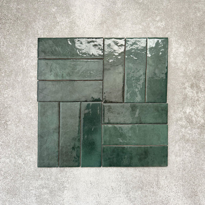 Flat lay of Zellige Royal Green Brick 6.5 × 20cm Moroccan-style ceramic tiles in rich deep green tones, arranged in a basketweave layout on a textured grey background, showcasing the glossy glaze, natural tonal variation and handcrafted zellige-effect finish.