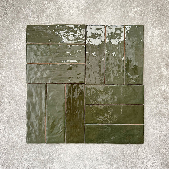 Flat lay of Zellige Botanical Green Brick 6.5 × 20cm Moroccan-style ceramic tiles in deep botanical green tones, arranged in a basketweave layout on a textured grey background, showcasing the glossy glaze, natural tonal variation and handcrafted zellige-effect finish.