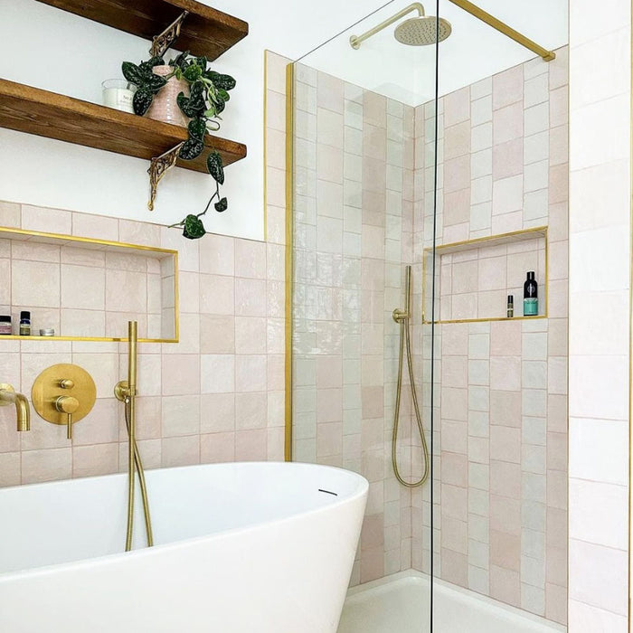 Bathroom featuring Zellige Pink Mist Square 13.2 × 13.2cm Moroccan-style ceramic wall tiles in soft pale pink tones, laid in a neat grid around a freestanding bath and walk-in shower, paired with brass fittings, glass screens and open shelving for a calm, elegant zellige-effect finish.