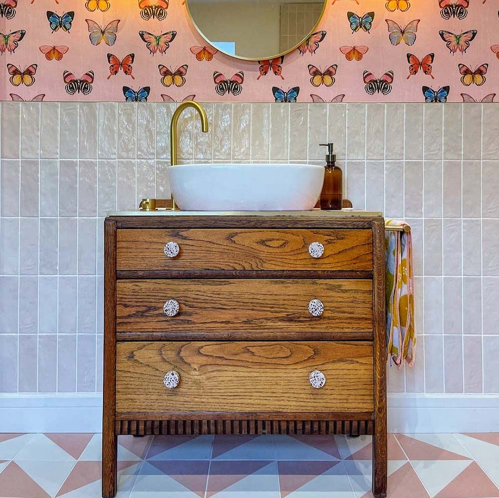 Bathroom vanity splashback featuring Zellige Pink Mist Brick 6.5 × 20cm Moroccan-style ceramic wall tiles in soft pale pink tones, laid in a vertical brick layout behind a white countertop basin with brass tap, paired with a wooden vanity unit, patterned wallpaper and geometric floor tiles to create a warm, playful and handcrafted zellige-effect finish.