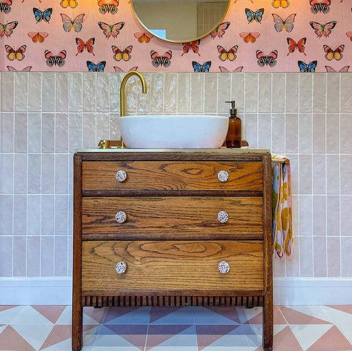 Bathroom vanity splashback featuring Zellige Pink Mist Brick 6.5 × 20cm Moroccan-style ceramic wall tiles in soft pale pink tones, laid in a vertical brick layout behind a white countertop basin with brass tap, paired with a wooden vanity unit, patterned wallpaper and geometric floor tiles to create a warm, playful and handcrafted zellige-effect finish.