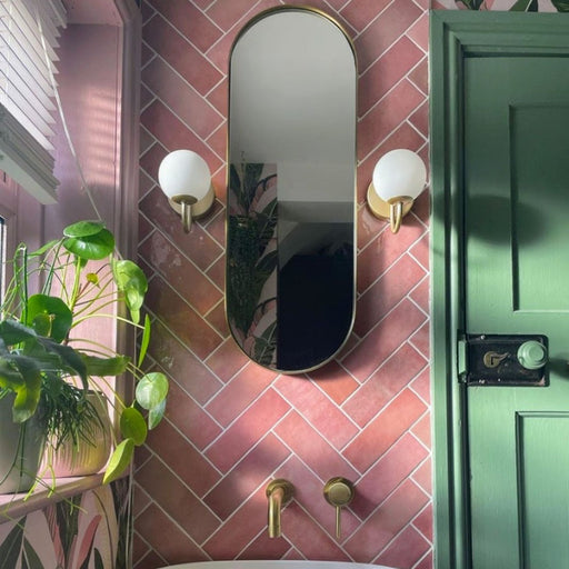 Bathroom wall featuring Zellige Rose Pink Brick 6.5 × 20cm Moroccan-style ceramic tiles, laid in a herringbone pattern with soft blush and dusty pink tones, paired with brass wall lights and taps, a green painted door and arched mirror to create a warm, characterful and handcrafted zellige-effect finish.
