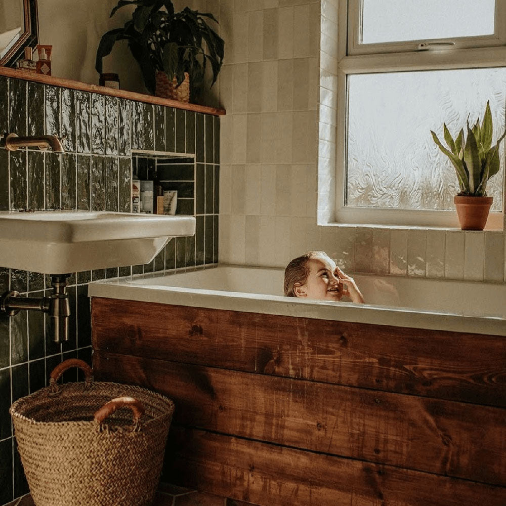 Bathroom featuring Zellige Botanical Green Brick 6.5 × 20cm Moroccan-style ceramic wall tiles in deep green tones, installed in a vertical brick layout around a built-in bath, paired with warm timber panelling, white sanitaryware and botanical accents for a rich, characterful zellige-effect finish.
