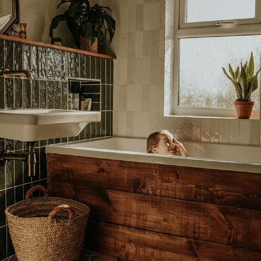 Bathroom featuring Zellige Botanical Green Brick 6.5 × 20cm Moroccan-style ceramic wall tiles in deep green tones, installed in a vertical brick layout around a built-in bath, paired with warm timber panelling, white sanitaryware and botanical accents for a rich, characterful zellige-effect finish.
