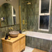 Bathroom featuring Zellige Malachite Green 6.5×20cm Moroccan-style ceramic brick tiles, installed on the walls around a bath and shower, paired with brushed brass fittings, a wooden vanity unit and a stone basin for a warm, spa-like finish.
