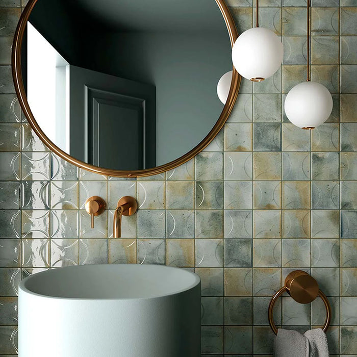 Bathroom wall featuring Saigon Celadon Green Decor Square Wall tiles in a Moroccan-inspired style, 10 × 10cm glossy ceramic tiles in soft green and blue-grey tones with a decorative relief pattern, paired with brass fittings and a round mirror.