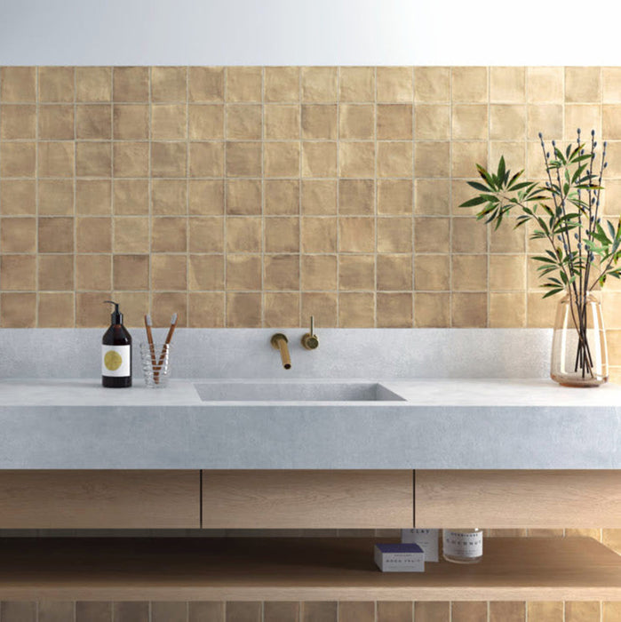 Bathroom splashback featuring Zellige Arena Marfil Square 10 × 10cm Moroccan-style ceramic wall tiles in warm beige tones, laid in a neat grid above a concrete-effect vanity with brass wall-mounted taps, with subtle colour variation and gently uneven edges adding an authentic handcrafted zellige-effect look.