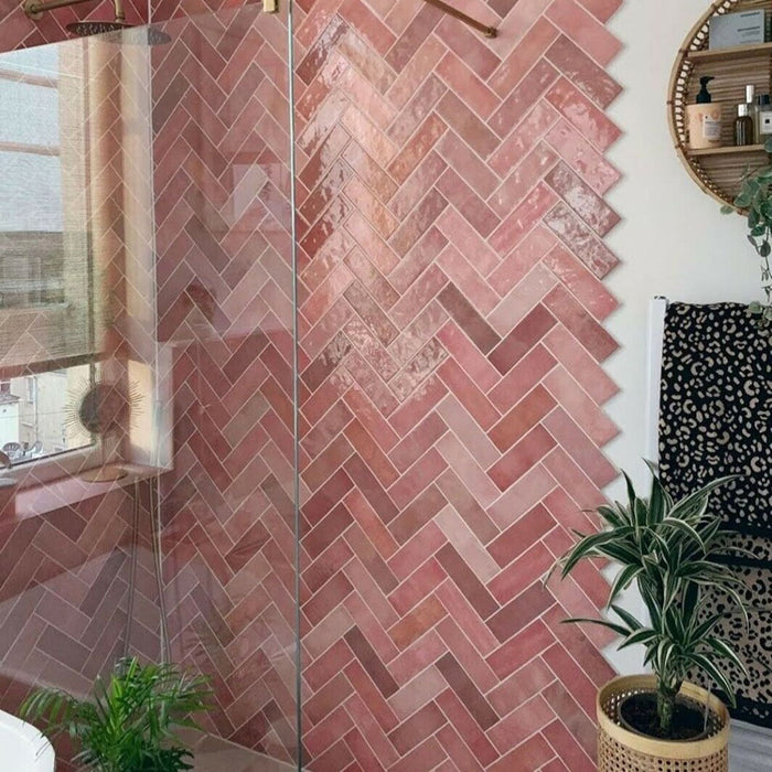 Bathroom wall featuring Zellige Rose Pink Brick 6.5 × 20cm Moroccan-style ceramic tiles, laid in a herringbone pattern with soft blush and dusty pink tones, paired with brass wall lights and taps, a green painted door and arched mirror to create a warm, characterful and handcrafted zellige-effect finish.