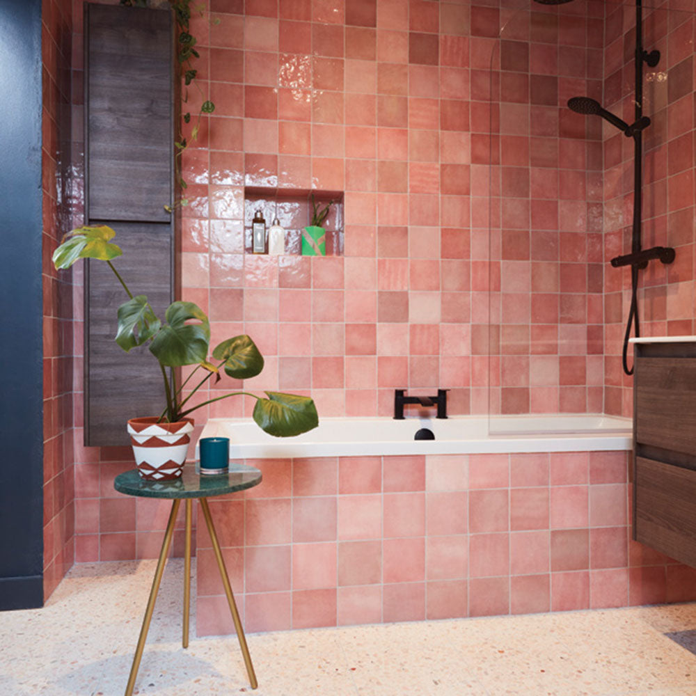 Bathroom featuring Zellige Rose Pink Square 13.2 × 13.2cm Moroccan-style ceramic wall tiles in soft blush and dusty pink tones, laid in a classic grid around a built-in bath and shower area, with a recessed shelf, black fittings and terrazzo-effect flooring highlighting the subtle tonal variation and handcrafted zellige-effect finish.