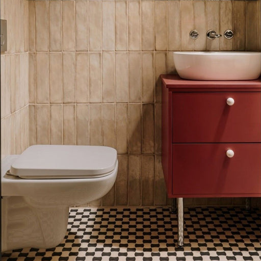 Bathroom featuring Zellige Arena Marfil Brick 6.5 × 20cm Moroccan-style ceramic wall tiles in warm beige tones, installed in a vertical brick layout behind a wall-hung toilet and red vanity unit, paired with patterned black and cream floor tiles and chrome fittings for a contemporary, characterful finish.