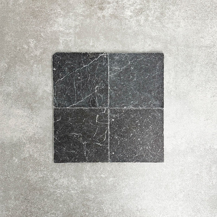 Four Fayrouz Black 15.2x15.2cm natural marble tiles with a tumbled finish arranged together, showcasing deep charcoal tones, natural veining and surface variation against a neutral textured background.
