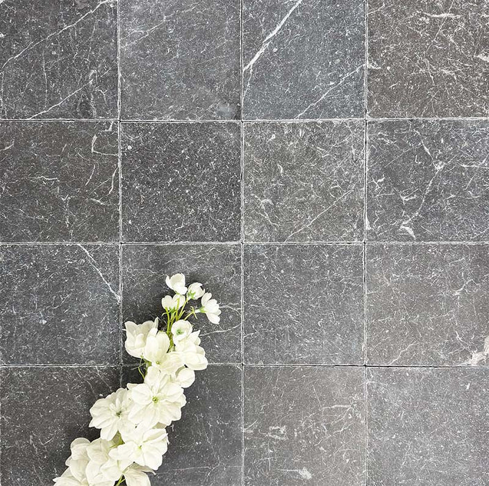 Flat lay of Fayrouz Black 15.2x15.2cm natural marble tiles with a tumbled finish, featuring deep charcoal tones and natural veining, styled with soft white flowers for contrast and texture.