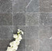 Flat lay of Fayrouz Black 15.2x15.2cm natural marble tiles with a tumbled finish, featuring deep charcoal tones and natural veining, styled with soft white flowers for contrast and texture.