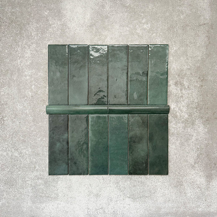 Flat lay showing Zellige Royal Green Brick 6.5 × 20cm Moroccan-style ceramic tiles paired with a matching Royal Green Bullnose 3 × 20cm trim tile in rich deep green tones, arranged on a textured grey background to highlight the glossy glaze, subtle tonal variation and handcrafted zellige-effect finish.