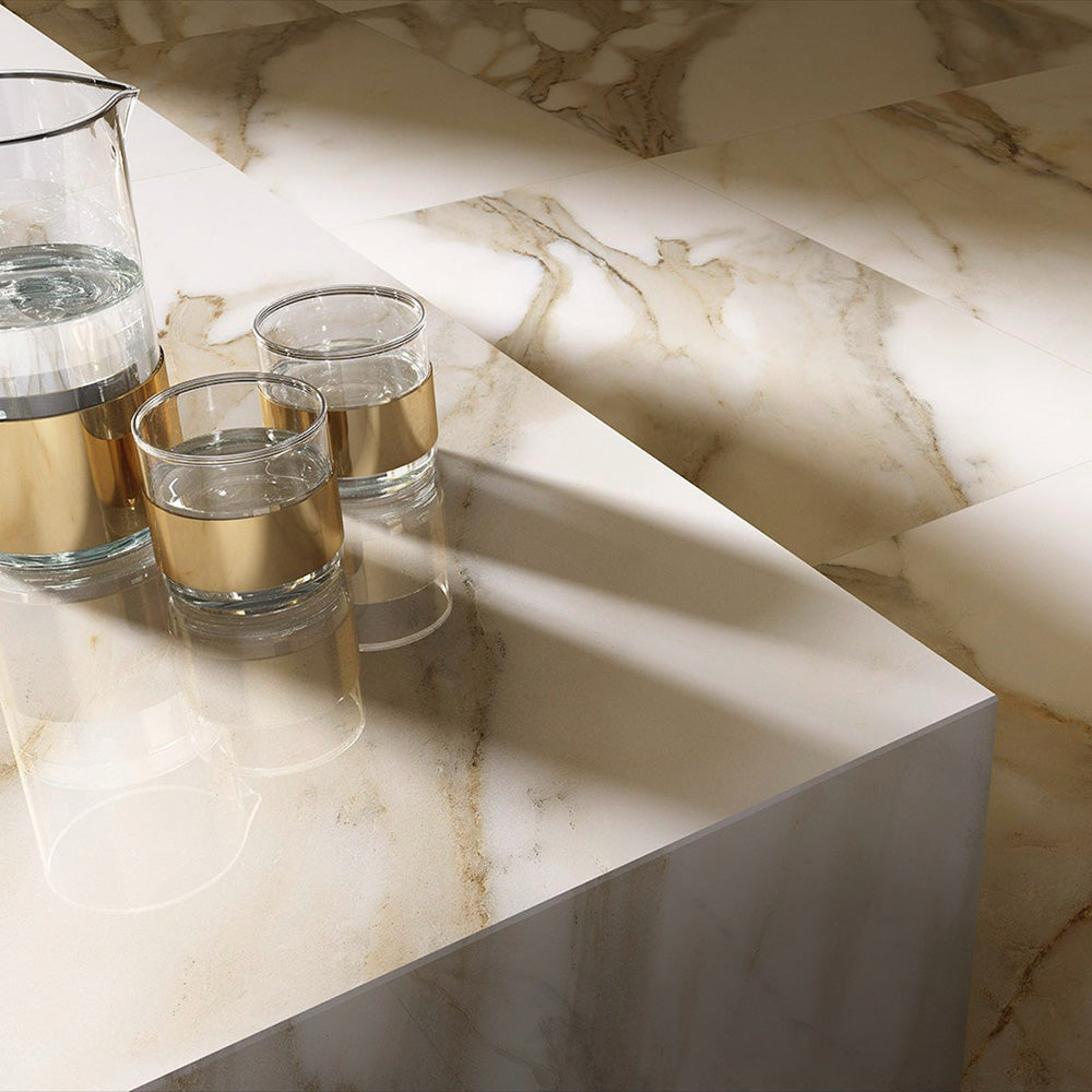 Close-up of Calacatta Oro Complement 60 × 60cm porcelain tiles, highlighting soft white tones with flowing gold veining, styled with reflective glassware to emphasise the polished finish and elegant marble effect.