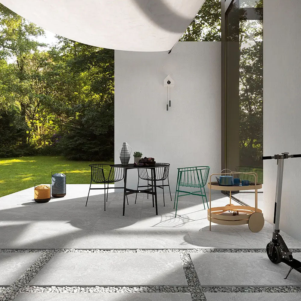 Outdoor patio setting featuring Comet Silver Stone 2cm 60 × 90cm porcelain tiles, laid in a modern grid with gravel joints. Soft silver-grey stone effect tones create a clean, contemporary look, complemented by minimalist furniture, greenery, and natural light.