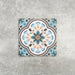 Cuba Bayamo Moroccan style patterned tile in a 20cm x 20cm format, featuring an ornate geometric floral design in soft blue, cream, teal and warm brown tones, displayed against a textured concrete-effect background.