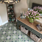 Hallway interior featuring Nantes Green Moroccan-style tiles in a 20x20cm format, laid as a decorative patterned floor. The soft green and white floral-inspired design adds a classic, timeless feel, paired with natural wood furniture, woven baskets and fresh flowers to create a warm, welcoming entrance space.