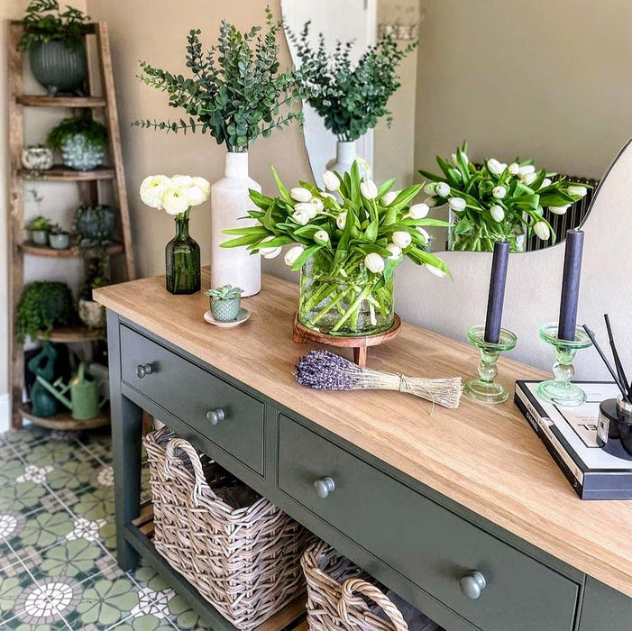 Hallway interior featuring Nantes Green Moroccan-style tiles in a 20x20cm format, laid as a decorative patterned floor. The soft green and white floral-inspired design adds a classic, timeless feel, paired with natural wood furniture, woven baskets and fresh flowers to create a warm, welcoming entrance space.