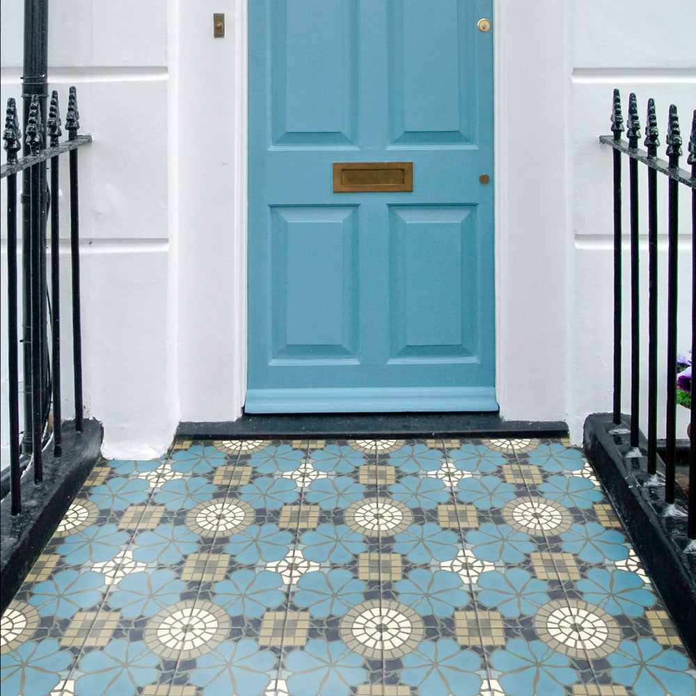 Nantes Azul Blue Moroccan-style tiles in a 20x20cm format, laid as a decorative outdoor entrance floor. The patterned tiles feature soft blue floral motifs with mosaic-style detailing in neutral and white tones, beautifully complementing the blue front door and creating a charming, characterful doorstep.